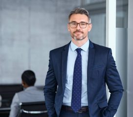 Portrait of handsome mid adult business man standing in modern office. Successful mature entrepreneur in formal clothing looking at camera with satisfaction. Confident man in suit with eyeglasses and beard standing with hands in pocket and looking at camera in office.